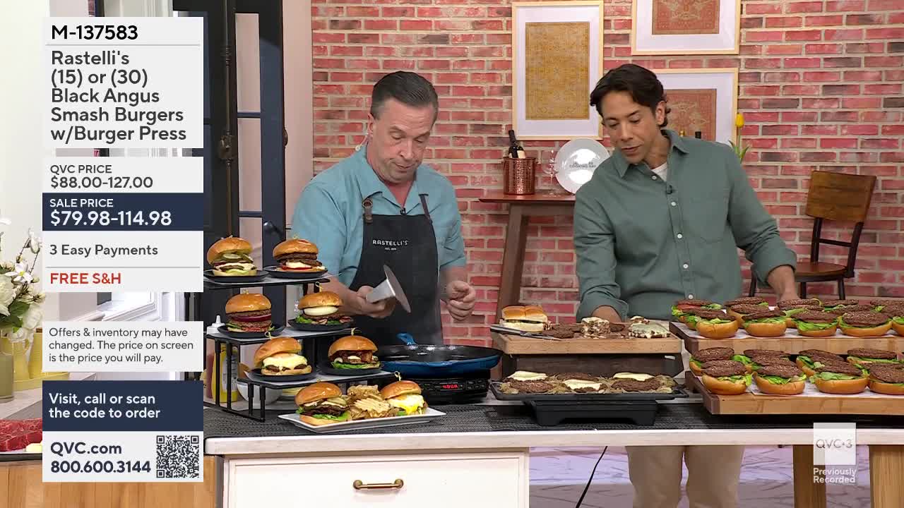 The man in the apron is pressing down on a burger in a pan. To his right, another man arranges rows of assembled burgers on a table.