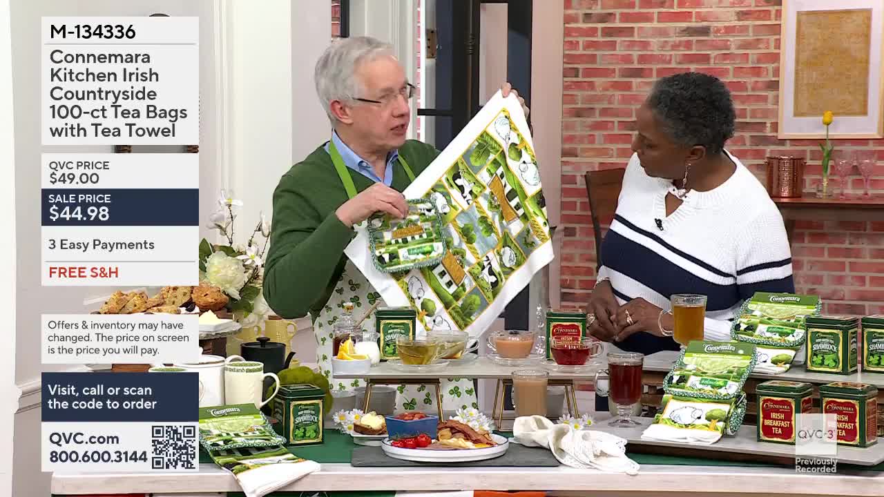 He holds up a tea towel printed with cows and green fields. On the table, several boxes of Connemara Irish tea sit next to cups of amber liquid.
