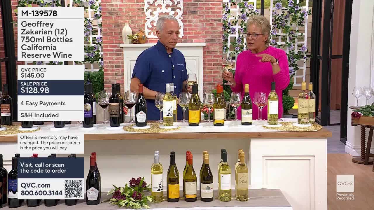 A man and woman stand behind a table displaying several bottles of wine. The woman holds a glass of white wine and gestures as she speaks.