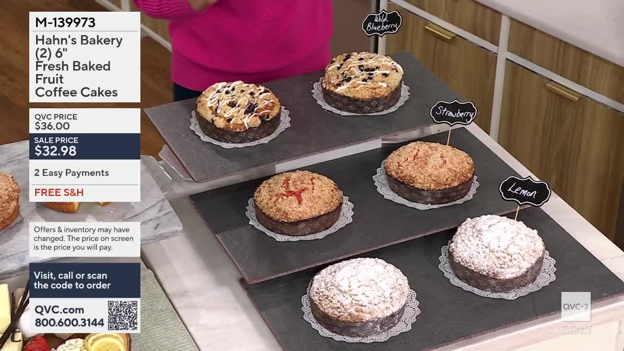 Six coffee cakes sit on tiered displays, each with a small chalkboard sign indicating its flavor. The blueberry cake on the top left has a crumb topping and visible berries. A cake dusted with powdered sugar sits on the bottom right.