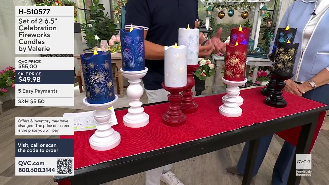 A man in a navy shirt gestures toward a row of decorative candles on a red runner. The candles, in blue, white, red, and black, are topped with flickering LED flames and feature firework designs.