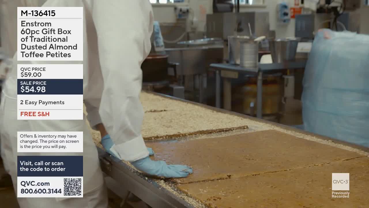 A worker in a white coat and blue gloves spreads a large sheet of toffee on a conveyor belt. The toffee is dusted with what looks like crushed almonds.