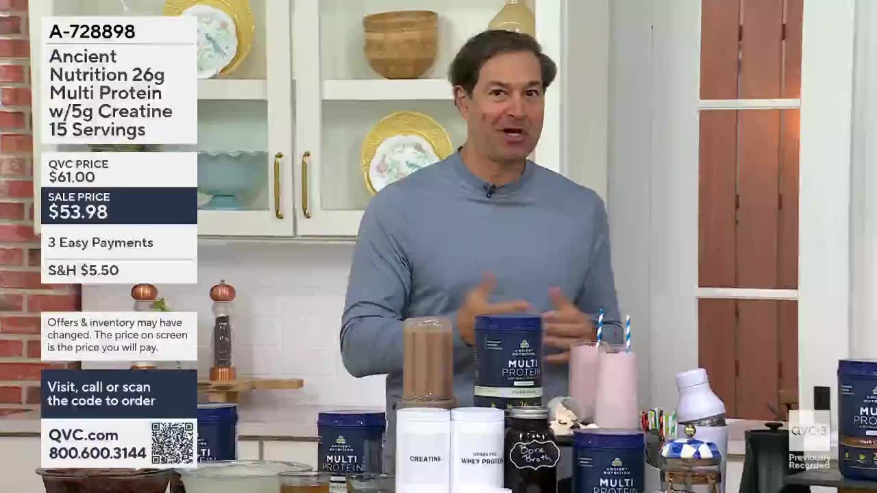 A man in a blue shirt gestures enthusiastically while standing behind a display of protein powder containers and drinks. Several blue and white containers of "Multi Protein" are arranged on a counter, along with two pink drinks in glasses.
