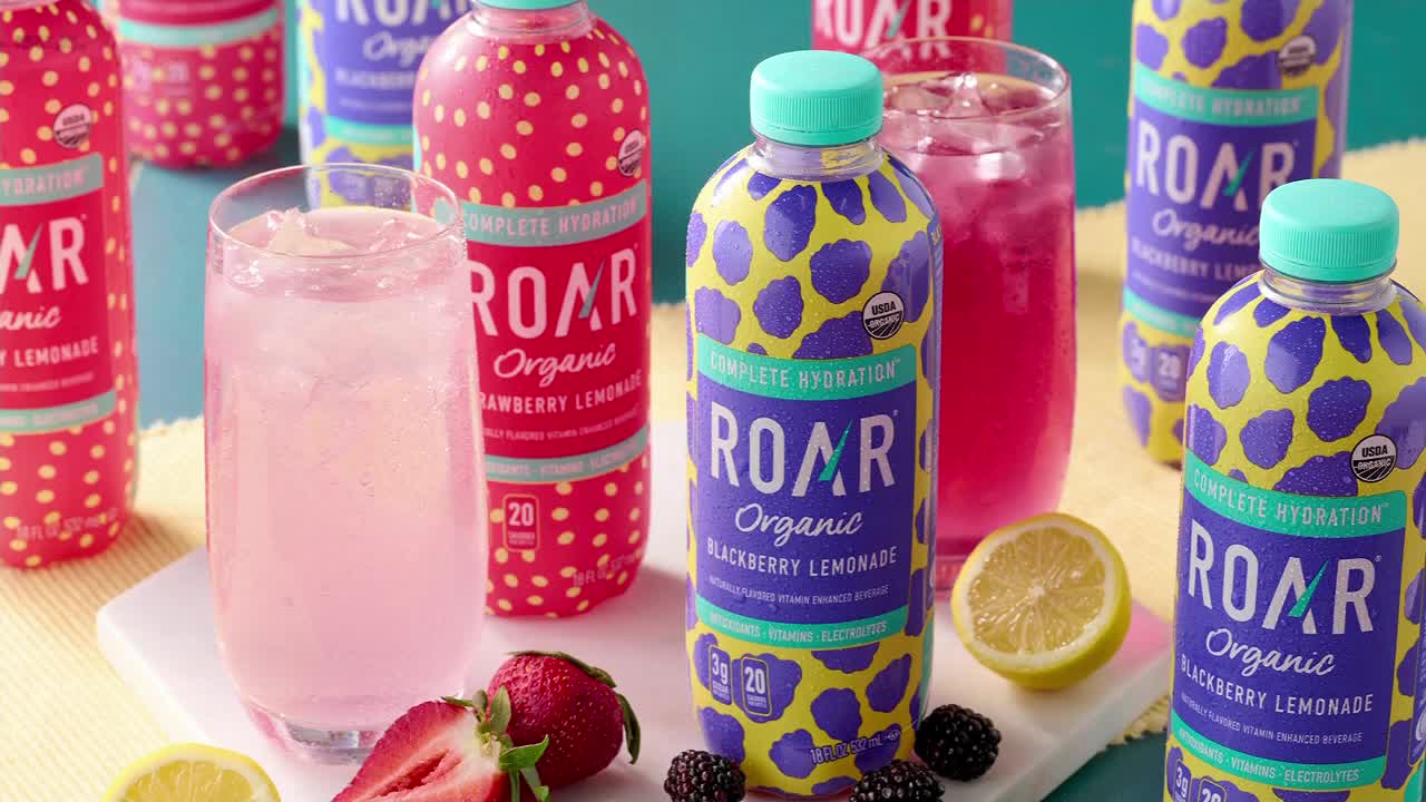 Bottles of ROAR Organic Strawberry Lemonade and Blackberry Lemonade stand ready. Glasses filled with pink and purple drinks sit beside fresh strawberries and blackberries.