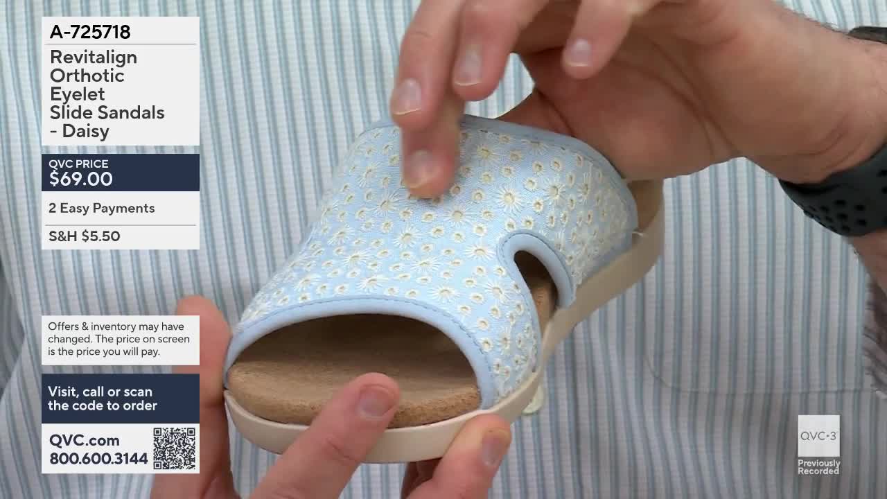 Fingers press into the light blue slide sandal, revealing its soft, patterned fabric. The sandal, featuring a delicate daisy print, is held up for display.