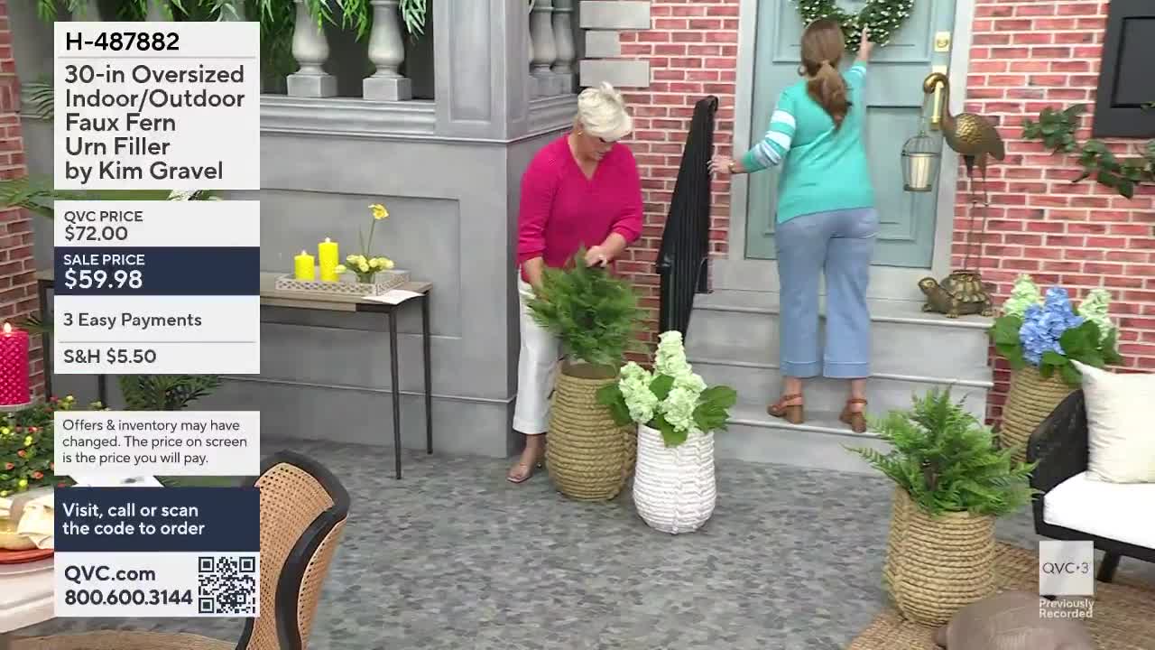 A woman in a pink shirt is placing a bushy green plant into a woven basket. Another woman, wearing a teal top, reaches up to hang a wreath on a blue door.