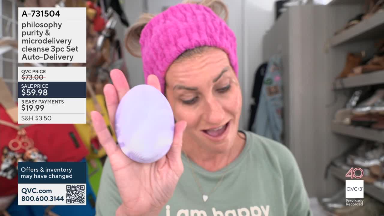 A woman in a pink headband holds up a lavender-colored oval device. She's on QVC3, showcasing a Philosophy skincare set.