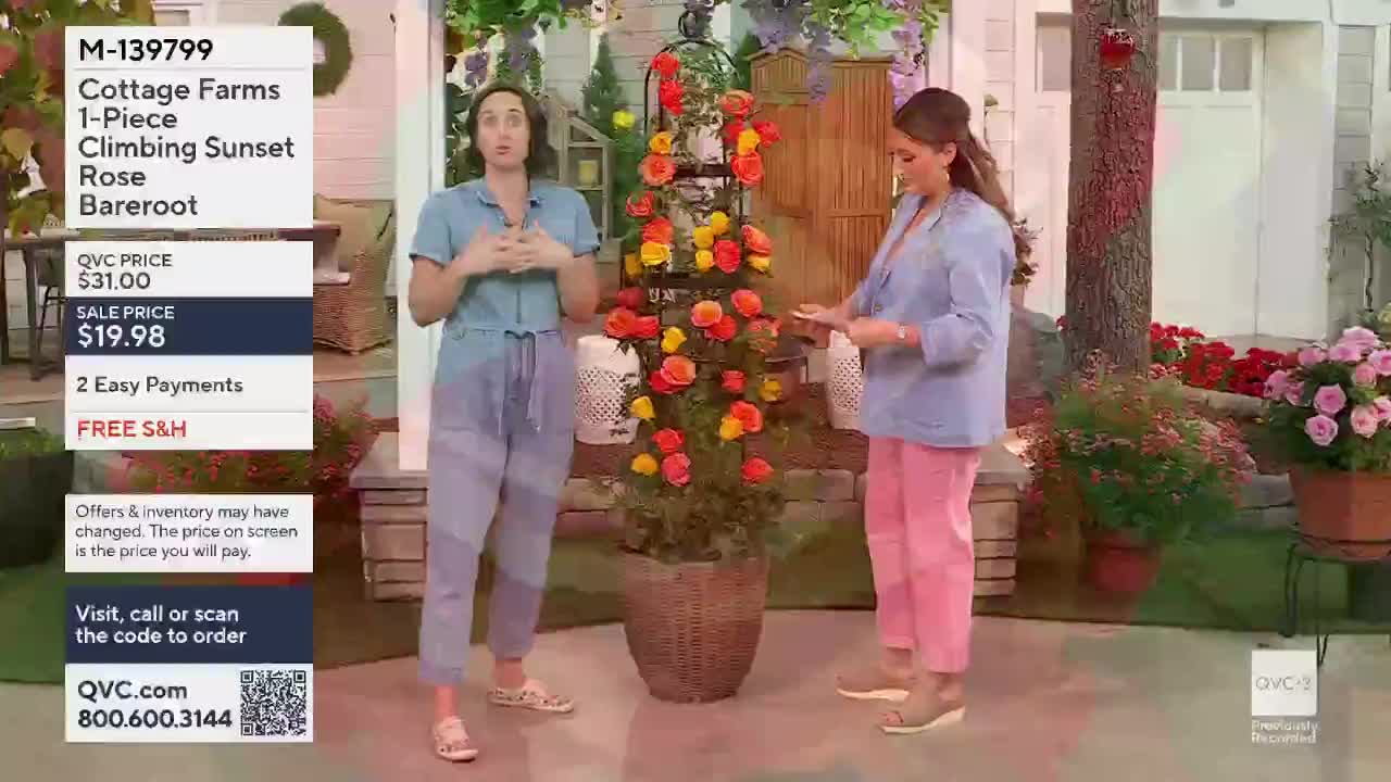 Two women stand beside a tall trellis covered in vibrant orange and yellow roses. The woman on the left gestures with her hands as she speaks, while the woman on the right holds a tablet.