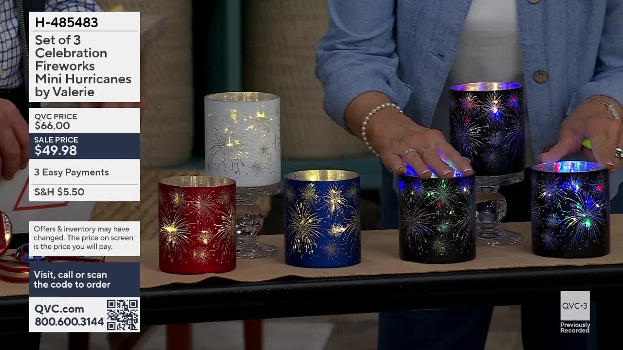 A woman's hands reach out, touching the tops of three black glass hurricane lanterns. Each lantern is decorated with a pattern of colorful fireworks that light up from within.