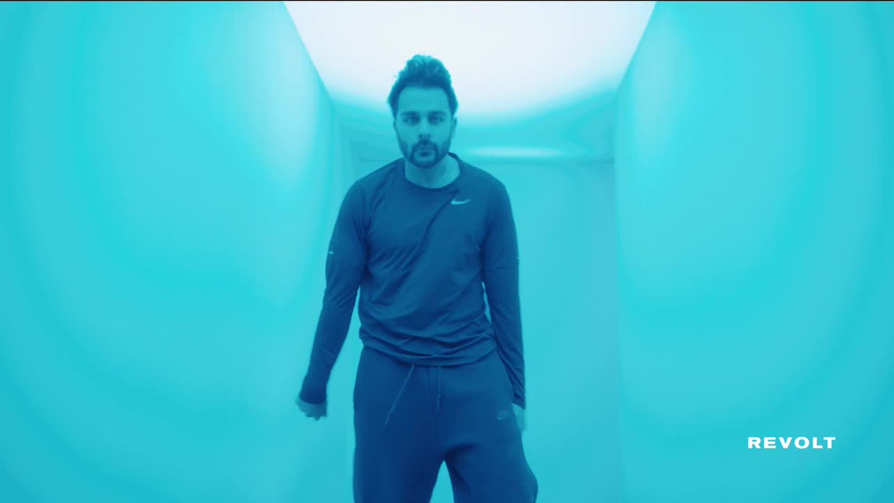 A man in workout clothes stands in a turquoise-lit corridor, looking directly at the camera. The Revolt logo is visible in the bottom right corner.
