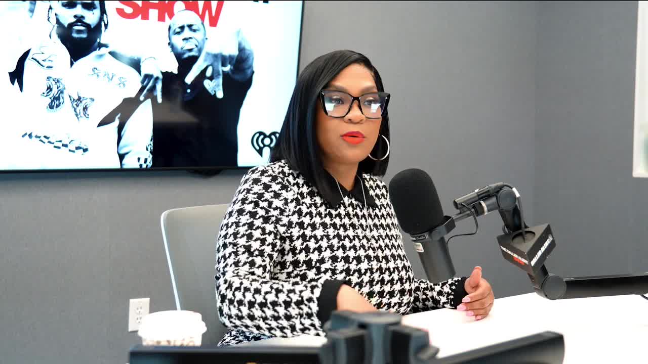 A woman in a houndstooth jacket sits at a desk, speaking into a microphone. Behind her, a large screen displays a black and white image of two men, and the iHeartRadio logo.
