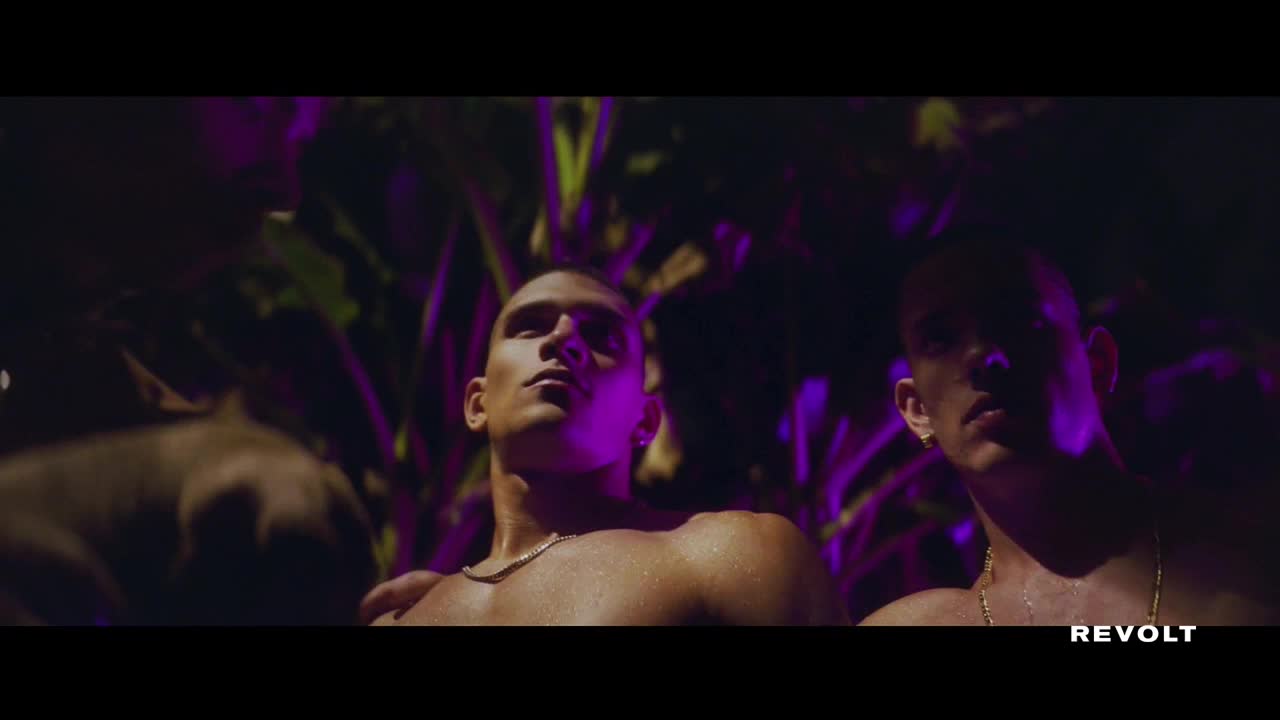 Three shirtless men stand close, illuminated by purple light. One looks up, another stares directly ahead, and the third is partially obscured.
