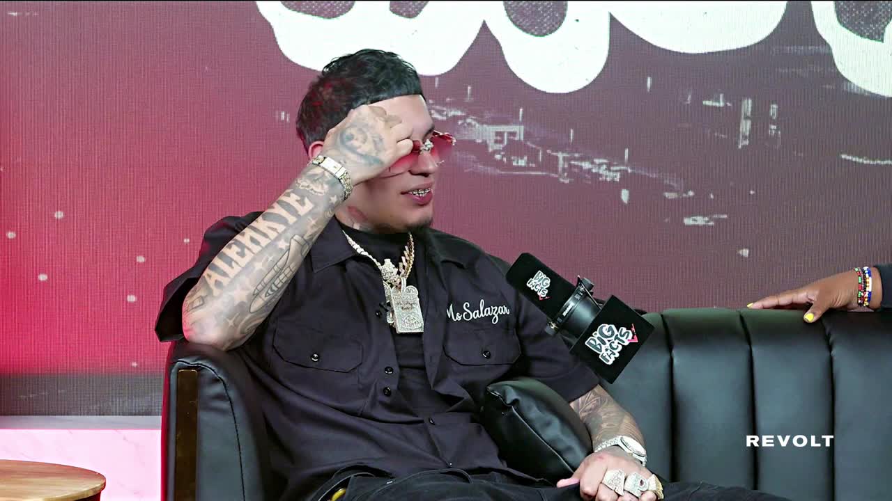 A man with heavily tattooed arms and a diamond-encrusted necklace sits on a black couch, hand raised to his sunglasses. He's being interviewed on a Revolt show, a microphone angled toward him.
