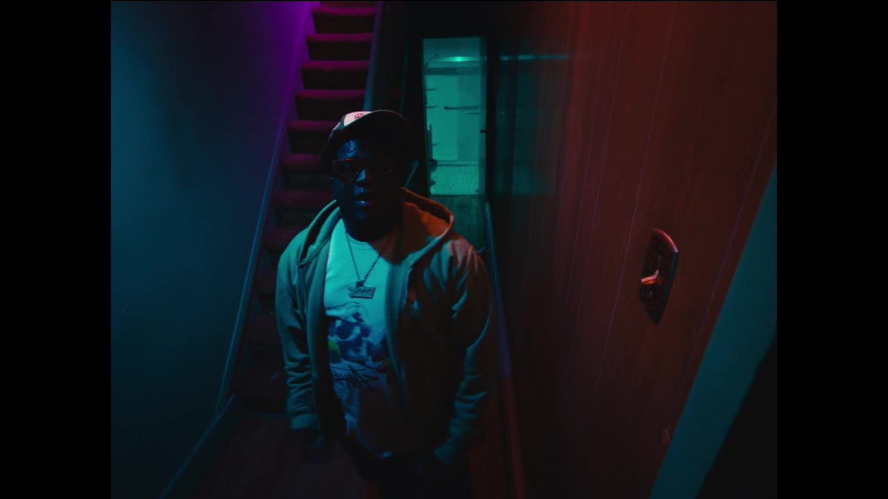 A man in a hoodie stands at the bottom of a dimly lit staircase. The walls are painted in shades of red and blue, casting an eerie glow on the scene.
