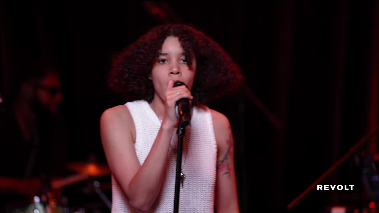 A woman with curly hair sings into a microphone, her face illuminated by a spotlight.  Behind her, the band is dimly visible in the red-tinged darkness.
