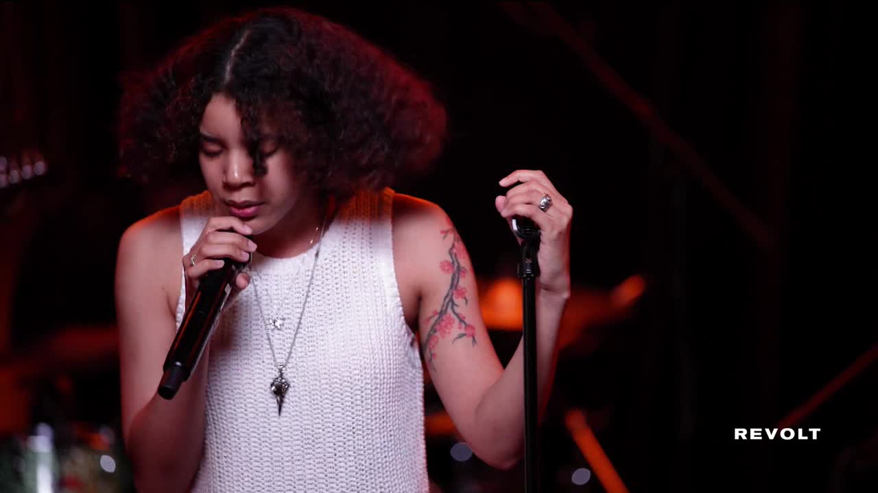 A woman with curly hair sings into a microphone, her gaze focused. The stage is dark, but a tattoo of a flowering branch is visible on her arm.
