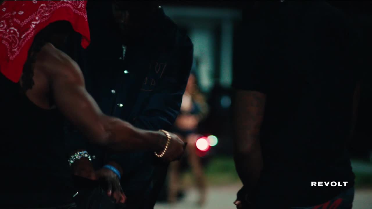 A man with a red bandana on his head is leaning in, his arm flexing.  He's next to another man wearing a dark shirt and a gold bracelet.
