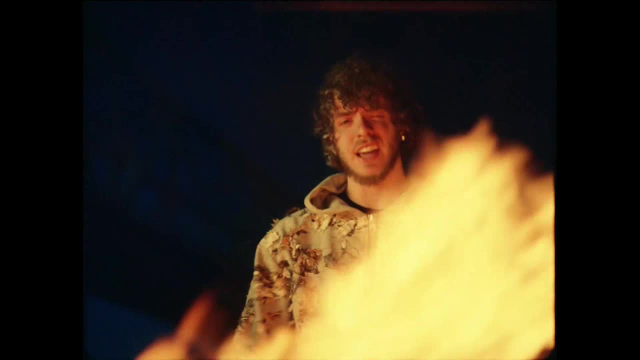 A man in a hoodie stands before a roaring fire, his face lit by the flames. He's singing, his mouth open as if he's belting out a song.
