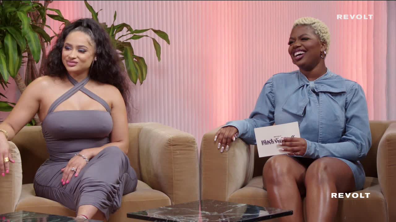 Two women are seated on plush armchairs, engaged in conversation. One holds a card with "Black Girl" visible, while the other smiles, her long, dark hair cascading over her shoulders.

