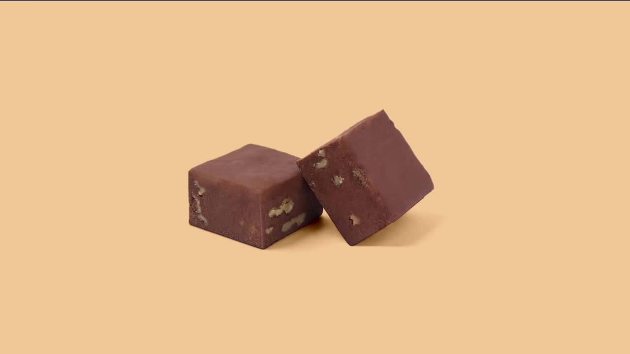 Two square pieces of fudge, speckled with nuts, sit on a plain background. One leans slightly against the other, casting a soft shadow.
