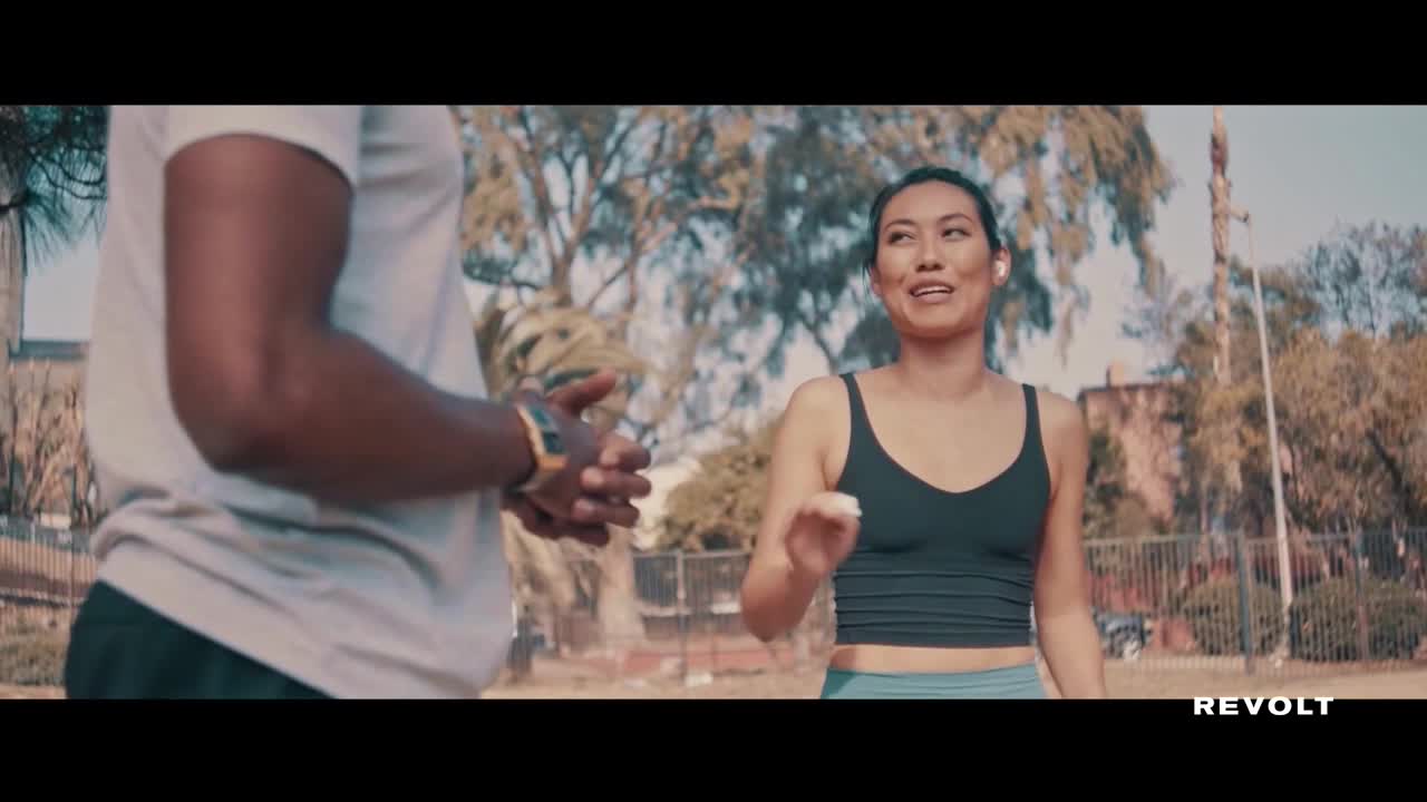 A woman in a black tank top is jogging, her face animated as she talks. Beside her, a man in a white shirt looks on, his hands clasped in front of him.
