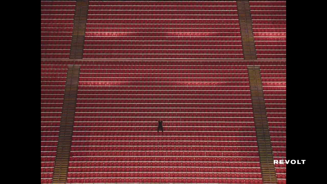 A lone figure stands in the middle of a stadium, dwarfed by the rows of empty red seats. The Revolt logo sits in the corner, a silent witness to the scene.
