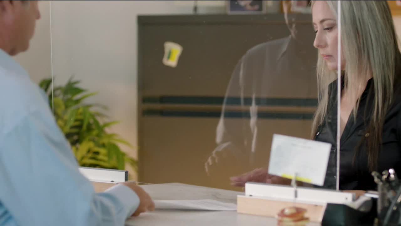 A woman behind a clear barrier is looking down at a document. Across from her, a man in a light blue shirt is also holding papers.
