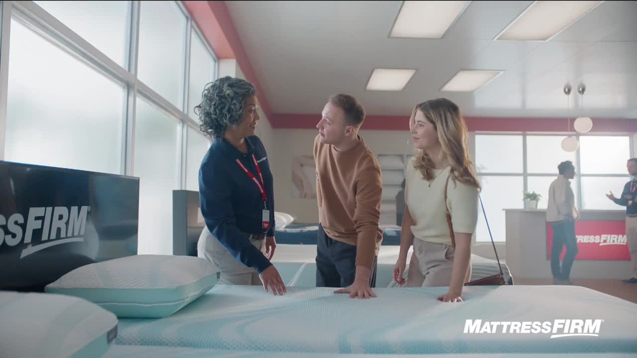 A salesperson gestures towards a mattress, explaining something to a couple. The man is leaning forward, while the woman looks on with a small smile.
