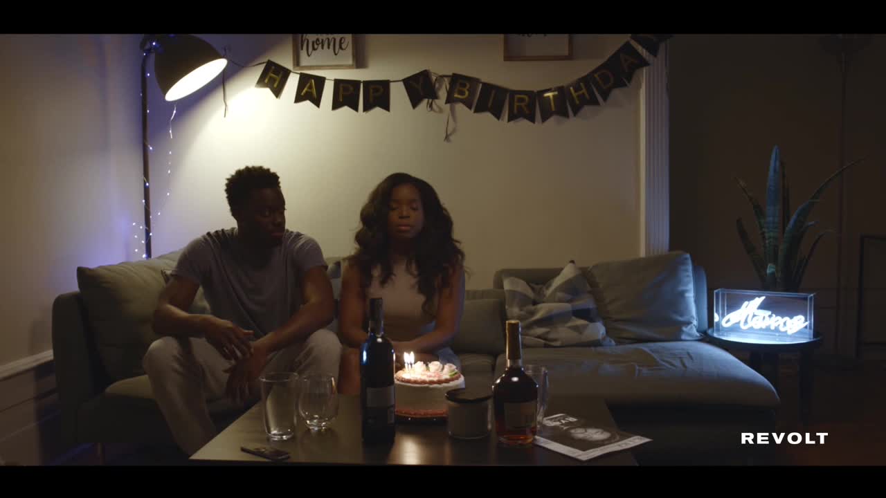 A couple sits on a couch, a small cake with lit candles between them. The woman closes her eyes, seemingly about to make a wish.
