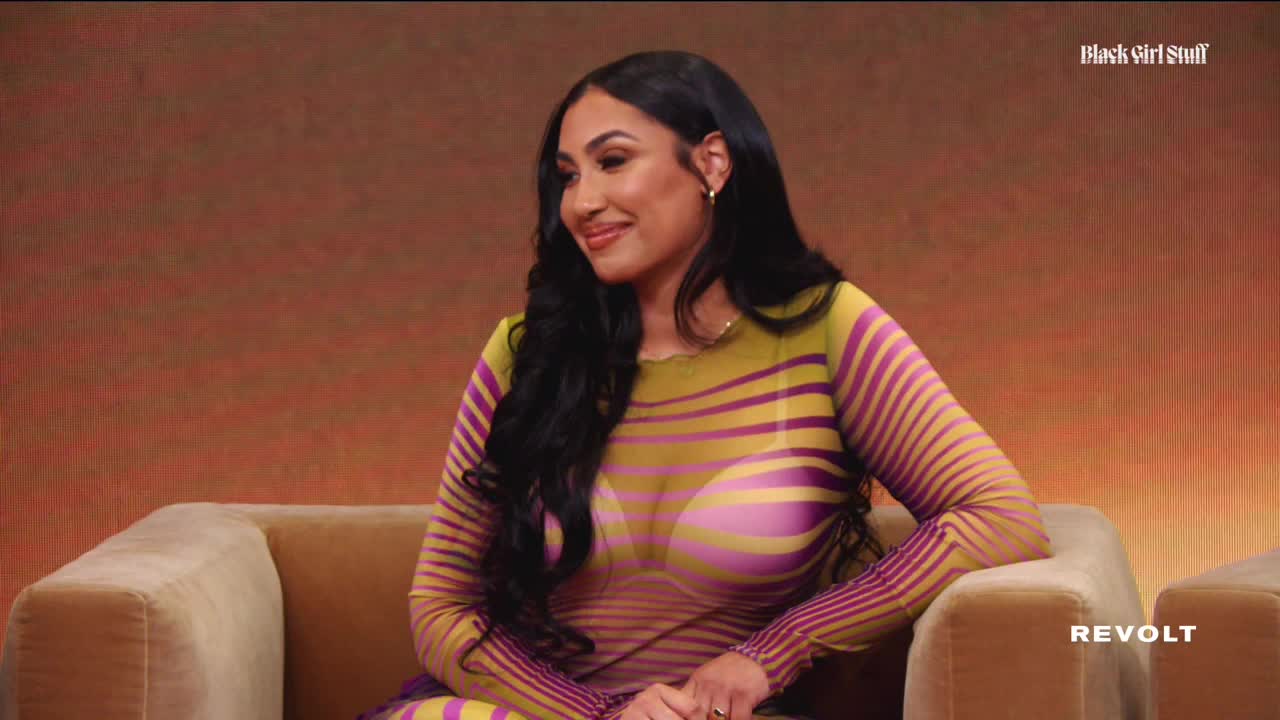 The woman in the striped dress is seated and smiling, looking slightly to her left. The REVOLT logo is visible in the bottom right corner.
