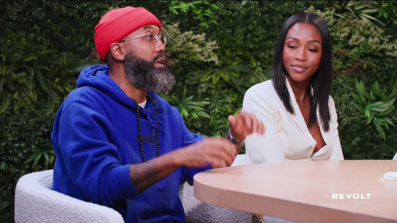 A man in a red beanie gestures with his hands, speaking animatedly. Beside him, a woman in a white blazer listens, her expression attentive.
