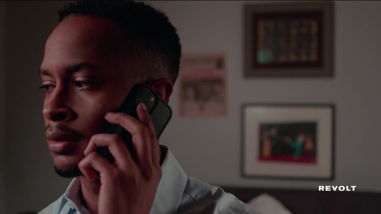 A man in a light blue shirt holds a phone to his ear, his gaze focused and intense. Behind him, framed pictures hang on the wall.
