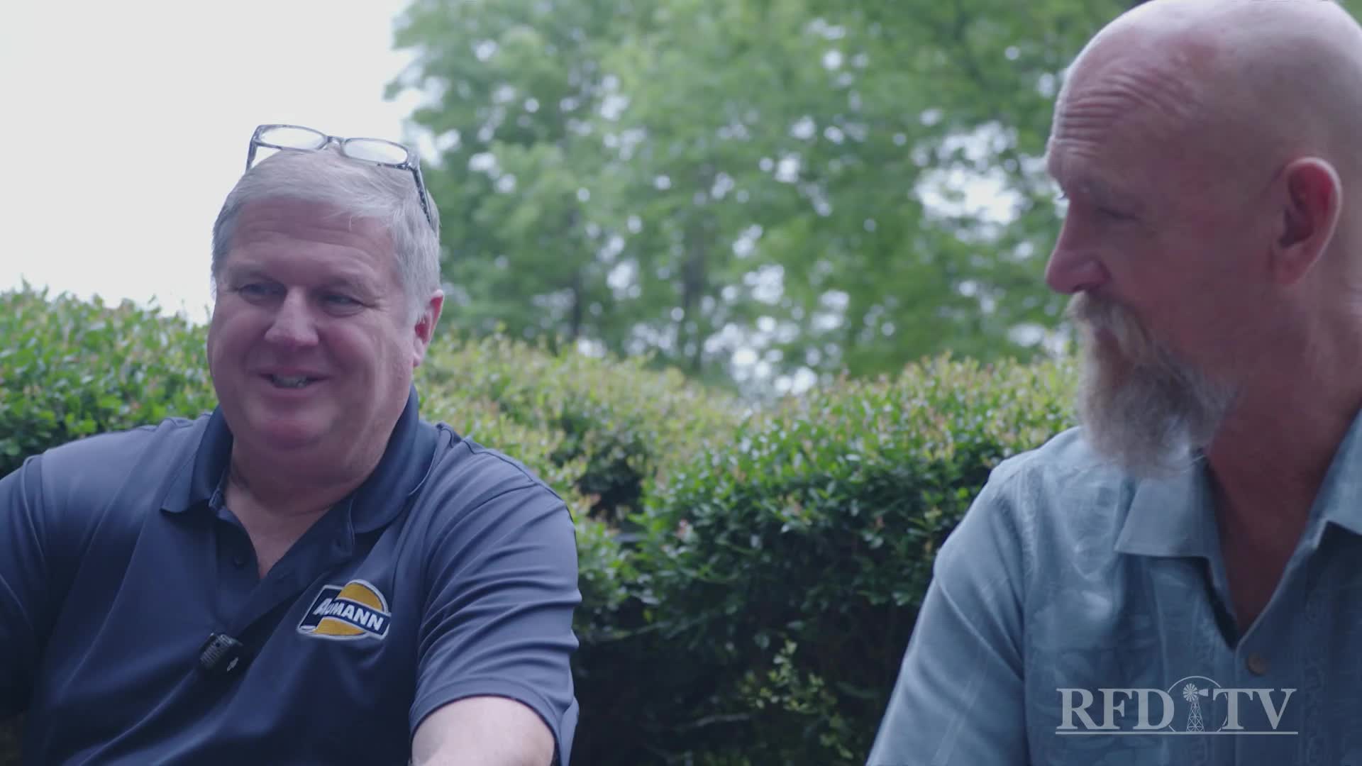 Two men are seated outdoors, talking. The man on the left, wearing a shirt with a logo, grins while the other listens intently.
