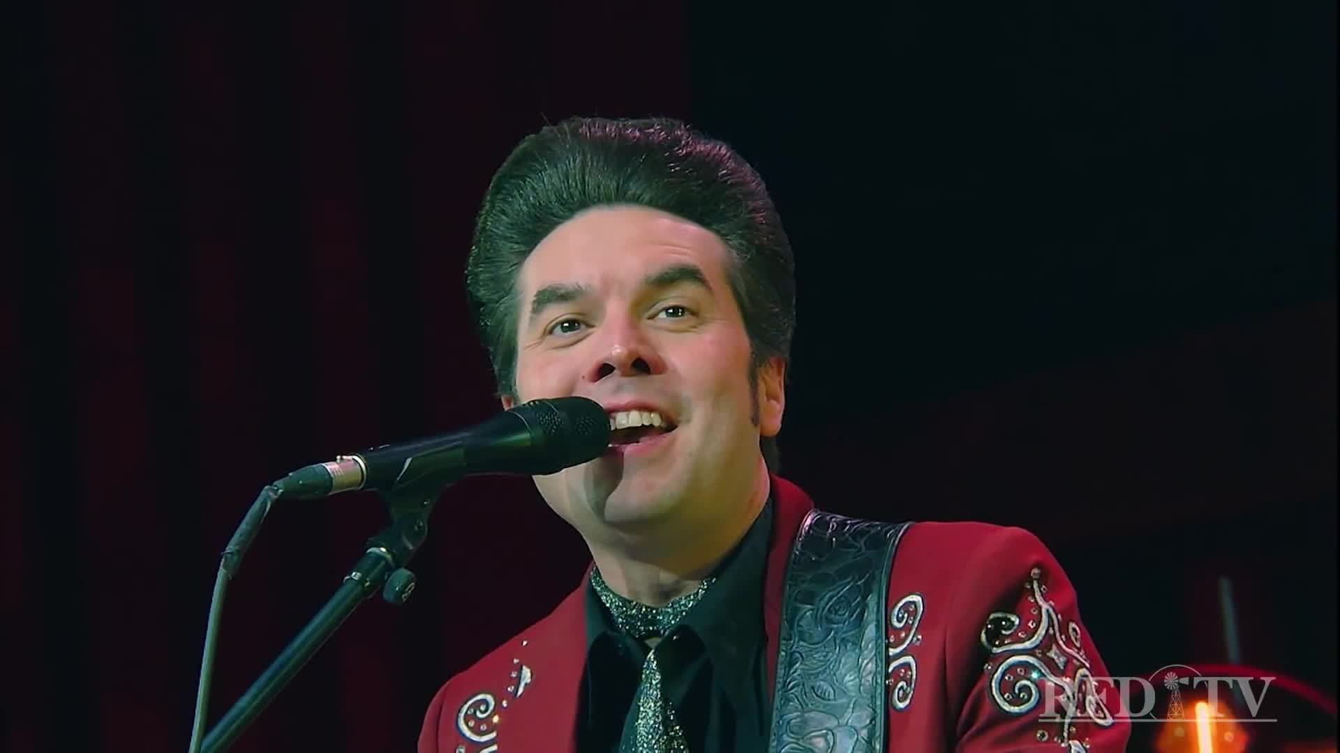 The singer is mid-song, grinning at the microphone, his hair styled high. He's wearing a red suit with ornate embroidery, and the RFD-TV logo is visible in the corner.
