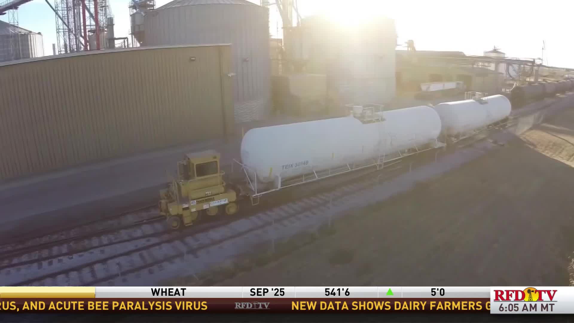 A small, yellow locomotive is pushing a line of white tanker cars along the tracks. The sun is bright, and the scene is being broadcast on RFD-TV.
