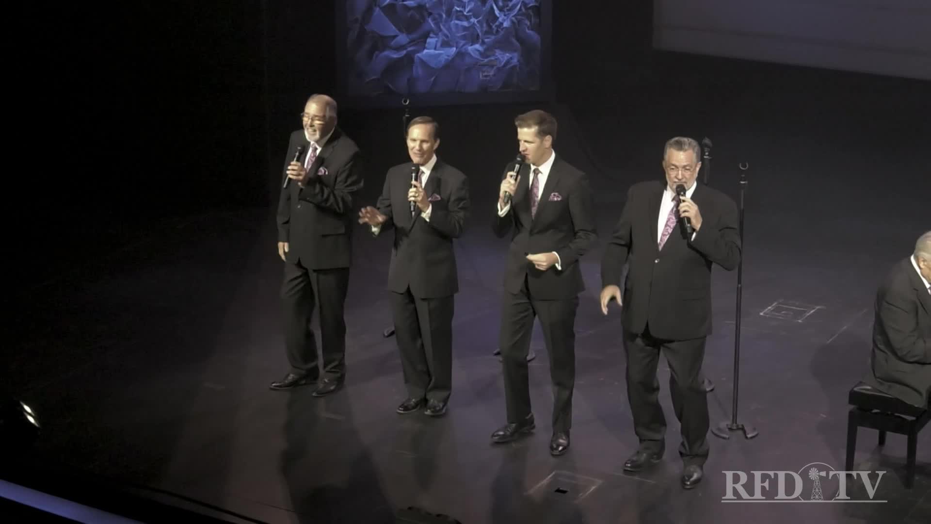 Four men in suits stand center stage, each holding a microphone as they address the audience. A fifth man, seated at a piano, is visible to the right.

