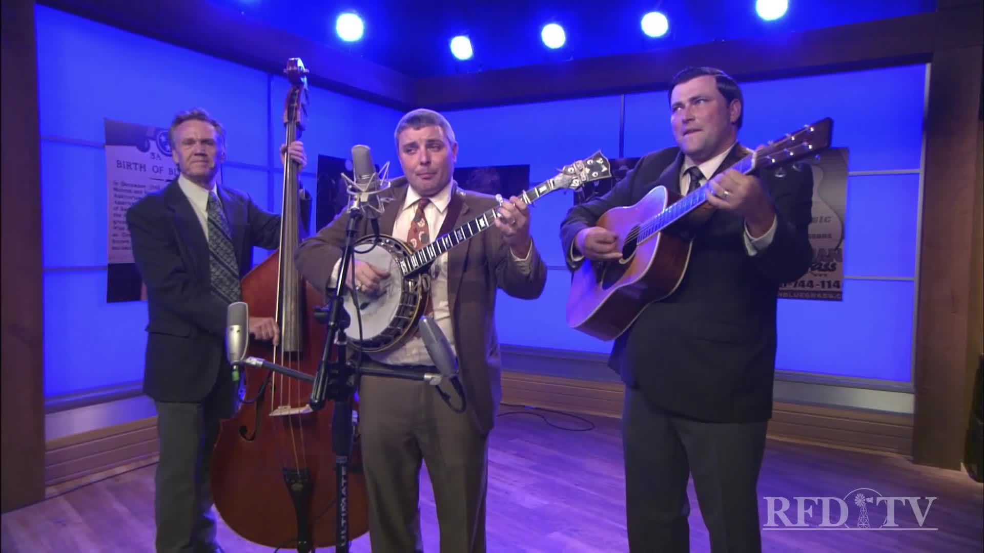 A trio of musicians are on stage, each playing their instruments with focus. The banjo player is singing into a microphone, while the guitarist and bassist provide accompaniment.
