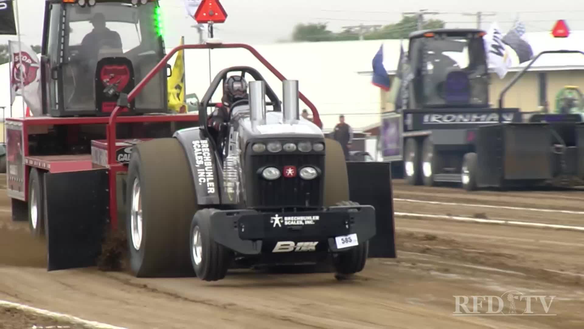 The black tractor, with its oversized tires, pulls a weighted sled down the dirt track.  A second truck, marked "IRONMAN," sits further down the track, as the event unfolds on RFD-TV.
