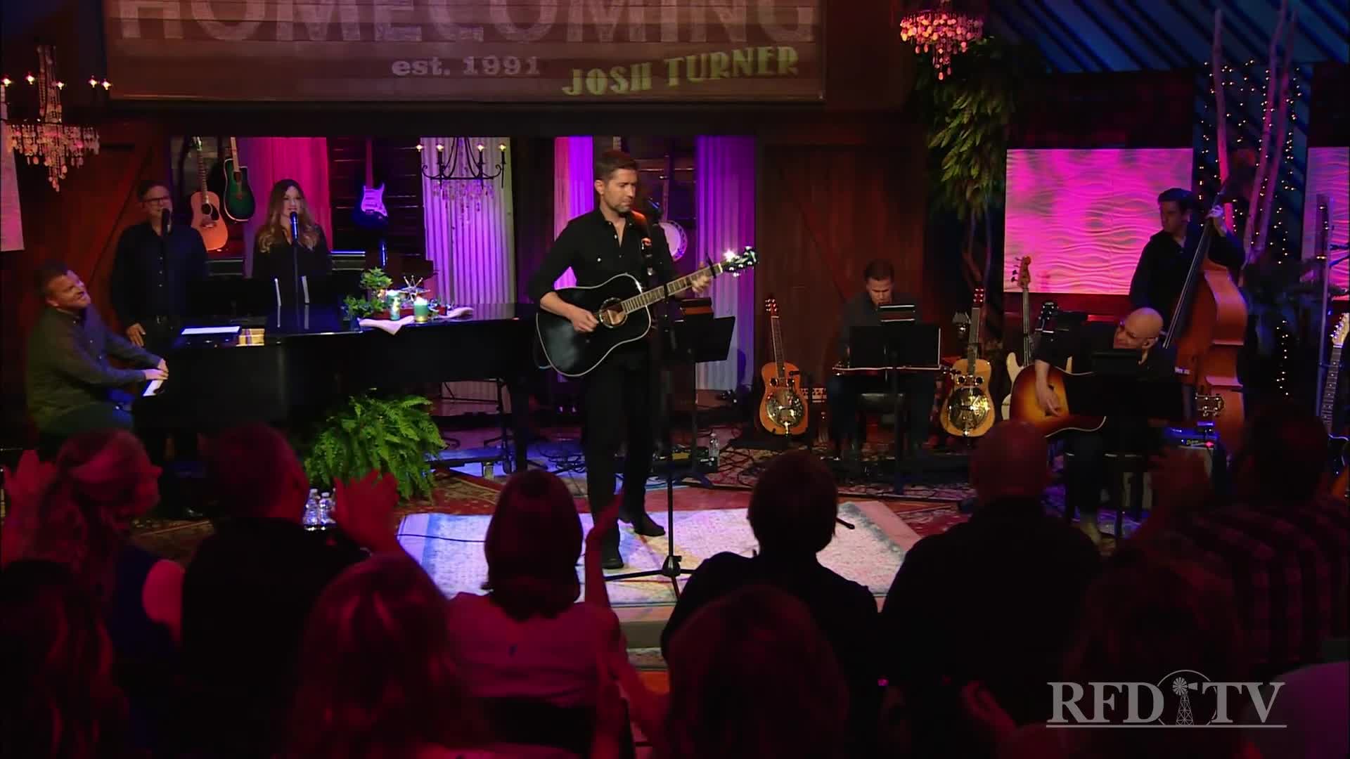 Josh Turner, dressed in black, strums his acoustic guitar center stage. A full band accompanies him, with a piano, bass, and backup singers.
