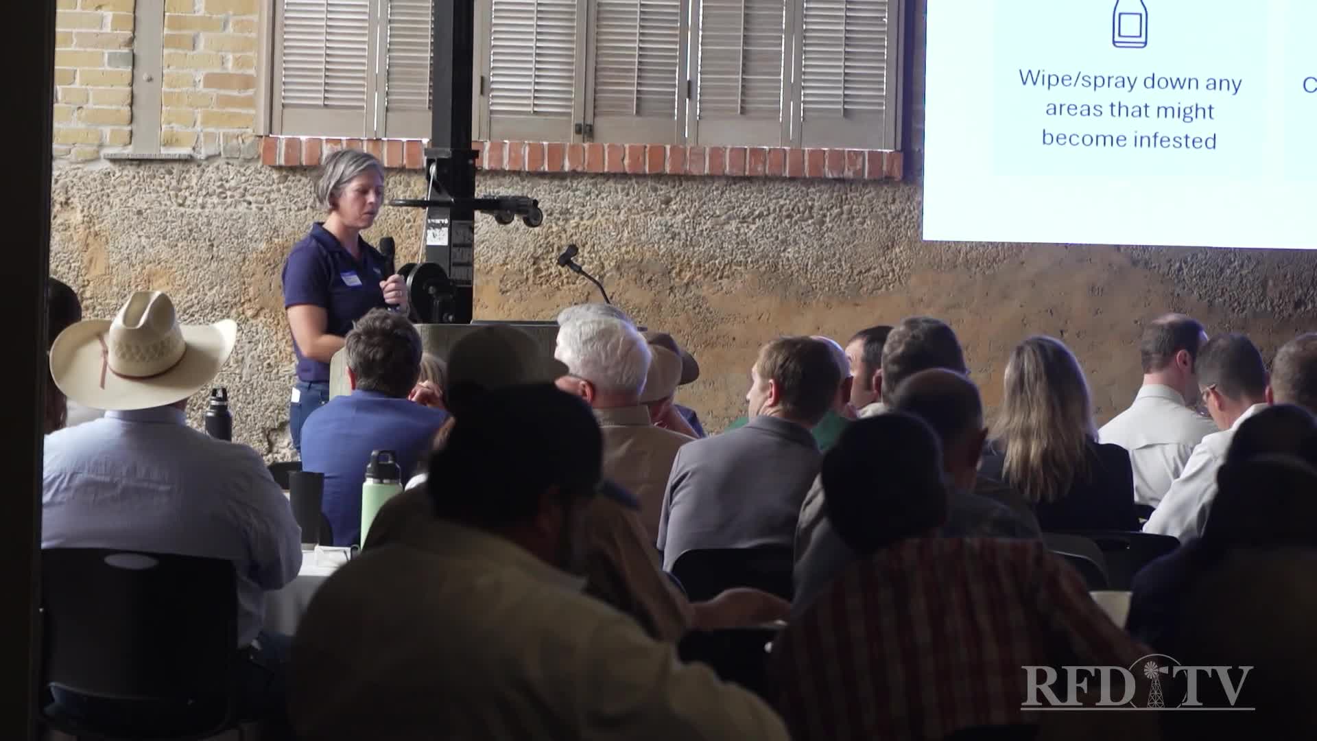 A woman with a microphone stands before a crowd, addressing them in a presentation. The RFD-TV logo is visible in the corner, and a screen behind her displays text about pest control.
