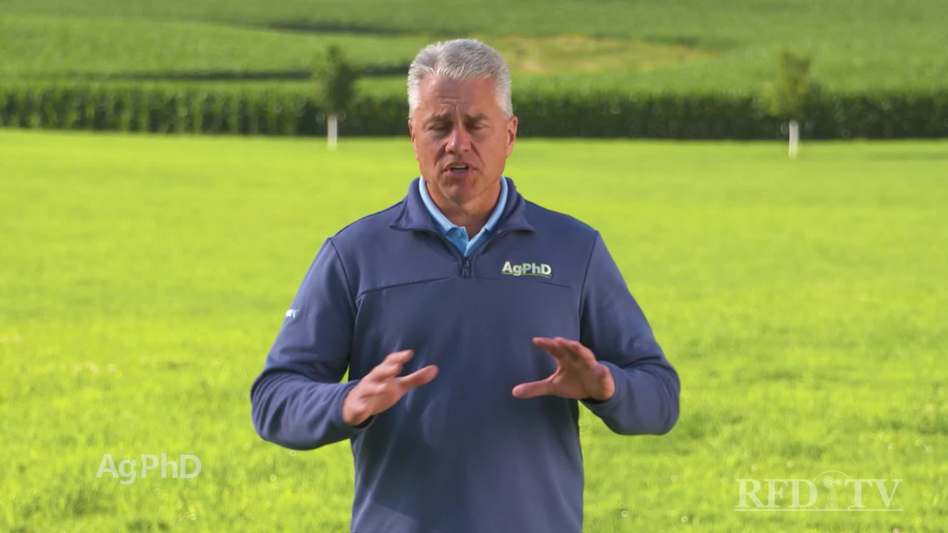 A man in a blue shirt stands in a field, gesturing with his hands. The RFD-TV logo is visible in the corner, and the background is a vast expanse of green crops.
