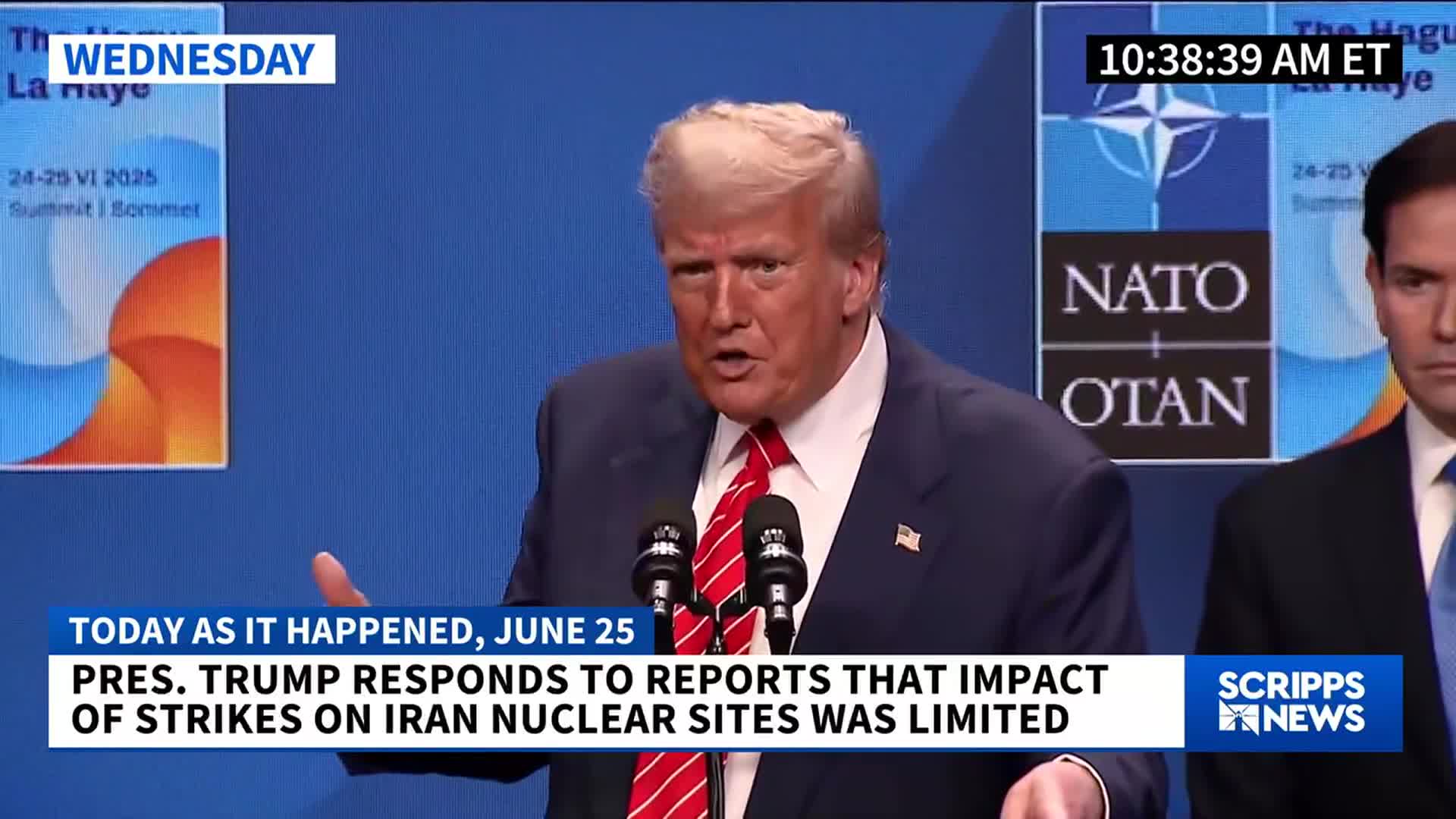 Donald Trump, wearing a blue suit and red tie, gestures emphatically as he speaks into two microphones. A Scripps News graphic at the bottom of the screen indicates the United States President is responding to reports about strikes on Iran.
