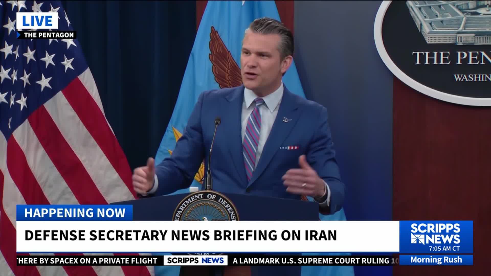 A man in a blue suit gestures with both hands from behind a podium at the Pentagon. The Scripps News graphic indicates a live briefing on Iran is underway.
