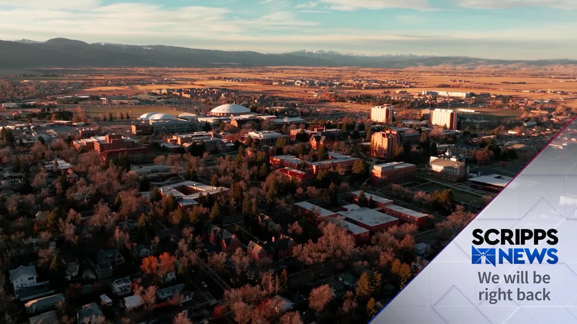 The sun is setting over a university campus, casting long shadows across the buildings. The Scripps News logo is in the corner, signaling a break in programming.
