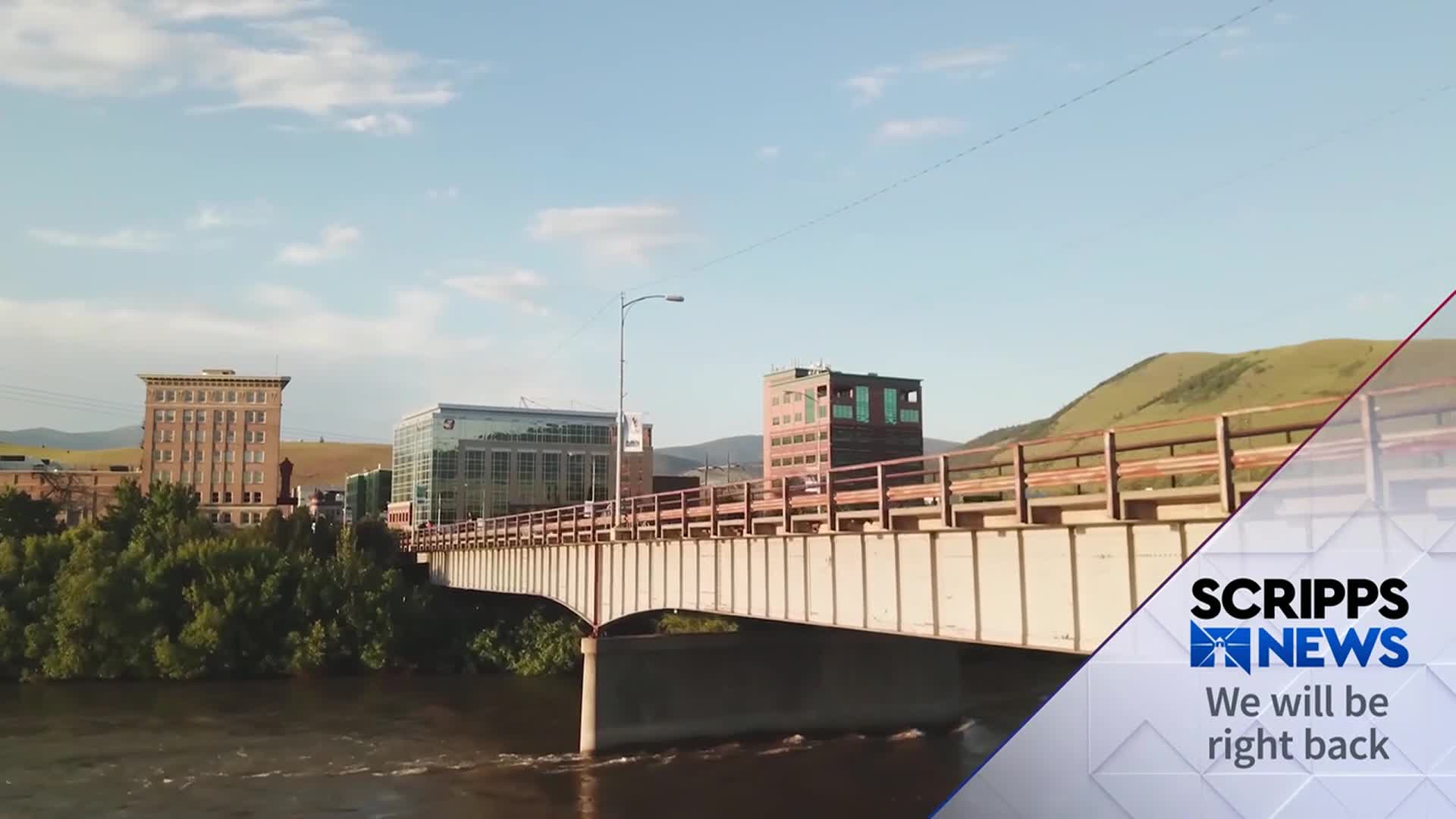 Water flows beneath a bridge spanning a river, with buildings lining the banks in the distance. The Scripps News logo in the corner indicates a break in the broadcast.
