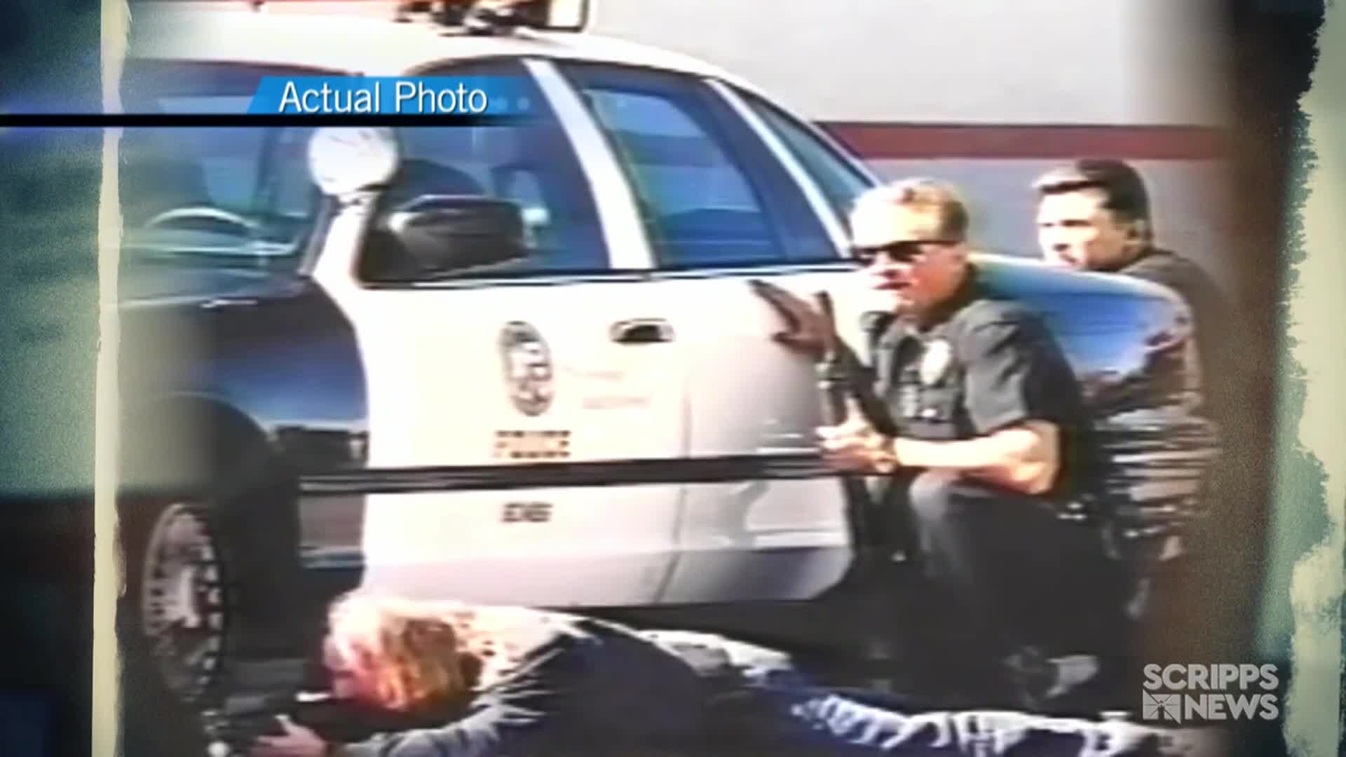 A person lies on the ground, aiming a long gun, while two police officers crouch near a patrol car. The "Actual Photo" is presented by Scripps News, and the scene appears to be a law enforcement situation in the United States.
