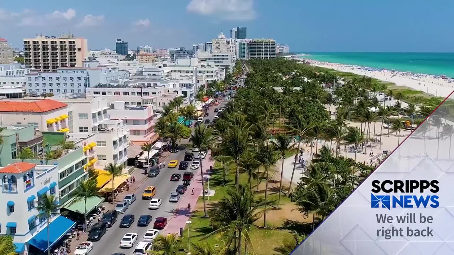 Cars crawl along a palm-lined boulevard in Miami, with the turquoise ocean and white sand beach visible in the distance. The Scripps News graphic in the corner indicates a break in the broadcast.
