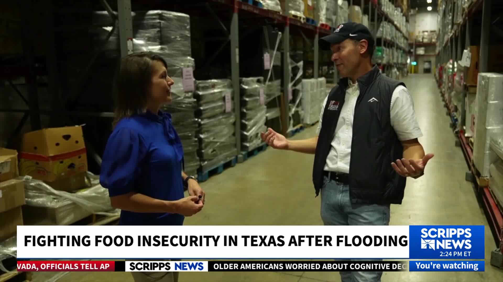 A man in a baseball cap gestures with both hands as he speaks to a woman in a blue blouse. They're standing in a warehouse filled with pallets of supplies, as Scripps News reports on food insecurity in Texas.
