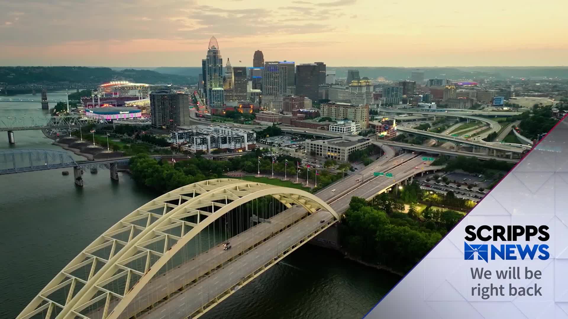 The sun is setting over a city, with a network of bridges and highways. A Scripps News graphic displays "We will be right back" at the bottom right.
