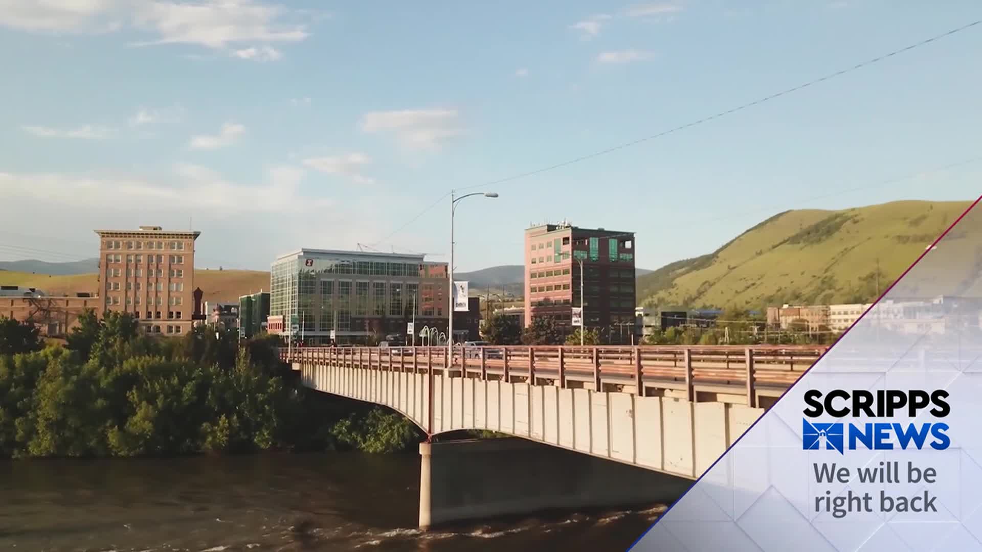 A bridge stretches across a river, connecting a city with buildings and a green hillside. The Scripps News logo indicates a break in programming.
